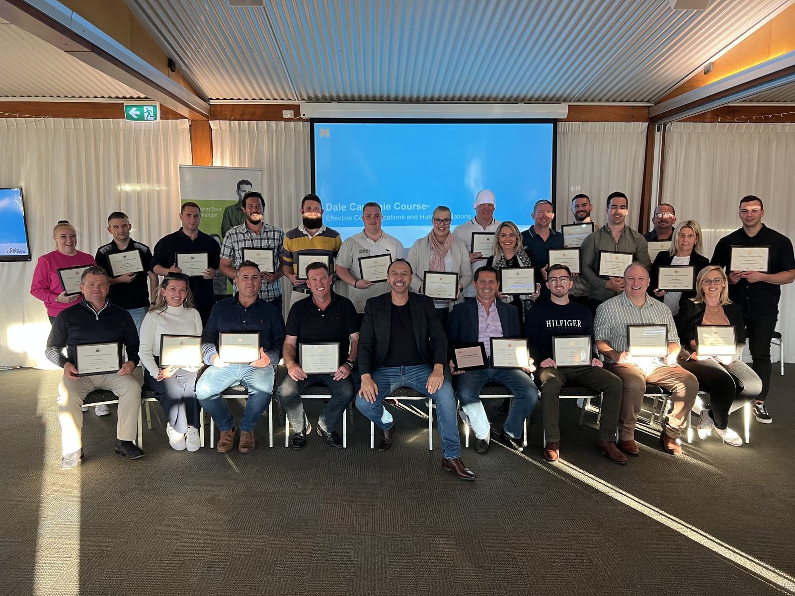 A graduating class of The Dale Carnegie Course holding their certificates of achievement in front of the course title on screen