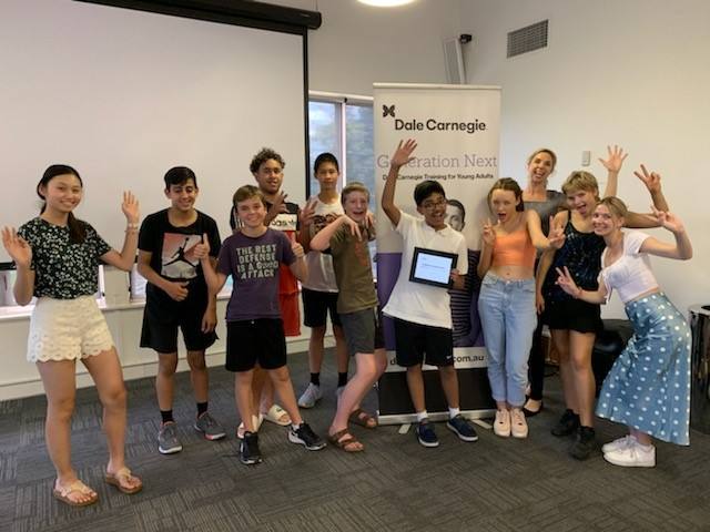Young adults from the Generation Next program waving beside a Dale Carnegie Generation Next banner, one holding a certificate