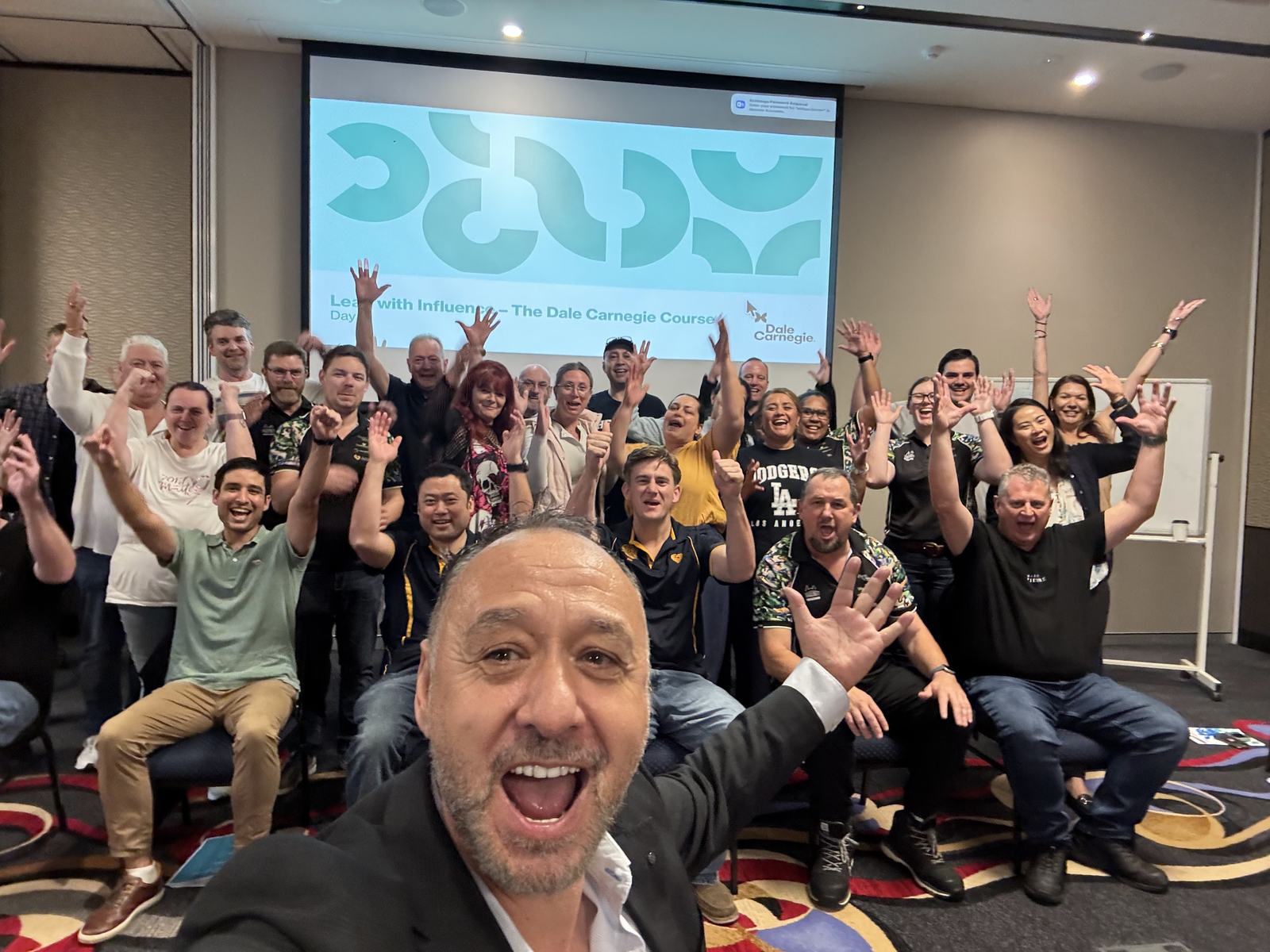 William Farmer taking a selfie with a cheering class in front of a screen reading Lead with Influence, The Dale Carnegie Course