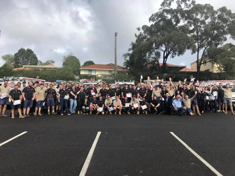 A large team in matching work uniforms holding certificates and cheering in an outdoor car park
