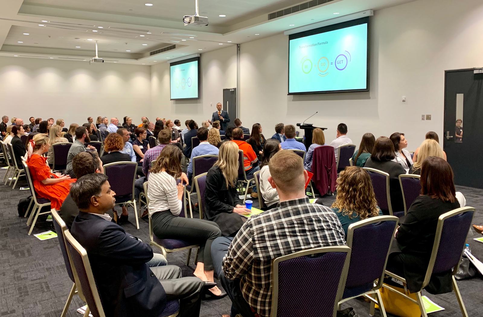 A packed workshop audience facing a presenter and screens showing the Transformation Formula, Be plus Do equals Get