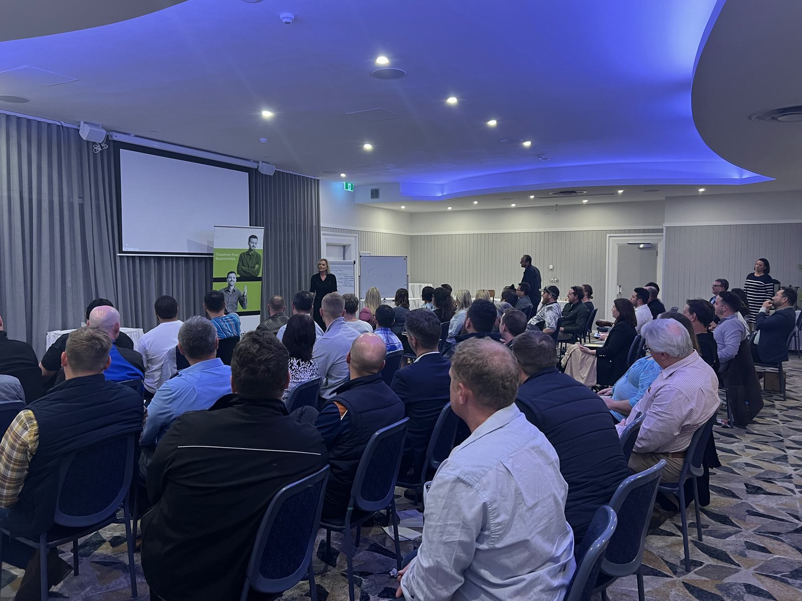 A full function room of people seated in rows, listening to a presenter beside a Dale Carnegie banner